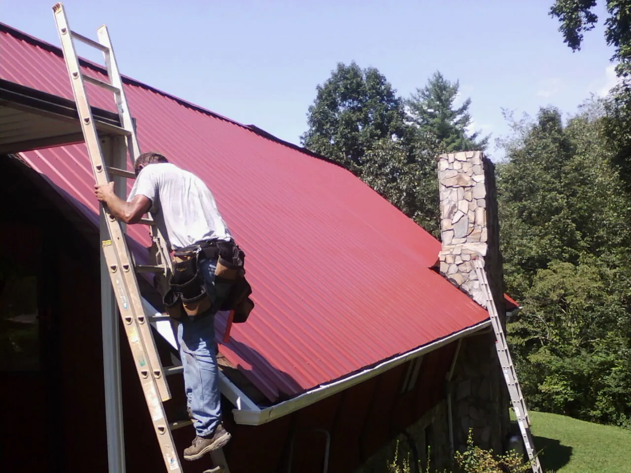 Roofer on a ladder beside a red metal roof and stone chimney — Soffit & Fascia Repair by Roofing Brothers in Wadena